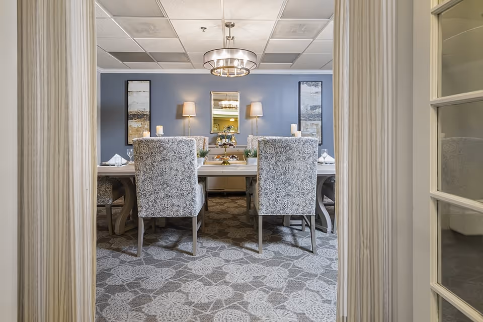 View through a doorway into a formal dining room with a large rectangular table surrounded by patterned upholstered chairs. The room has a blue accent wall with two framed artworks and two wall lamps on either side of a mirror. A modern chandelier hangs from the ceiling, and the floor is covered with a patterned carpet.