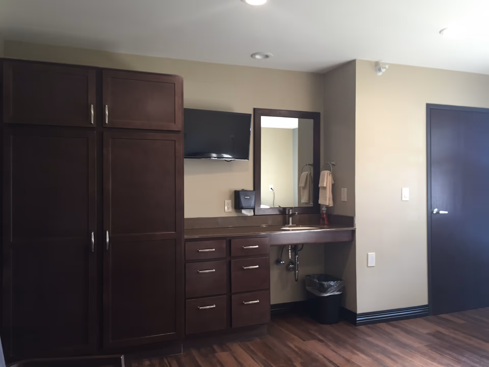 Interior view of a room with dark wood cabinets and drawers, a countertop with a sink, a mounted flat-screen TV, a mirror above the sink, beige walls, a closed dark wood door, and a trash bin underneath the counter.