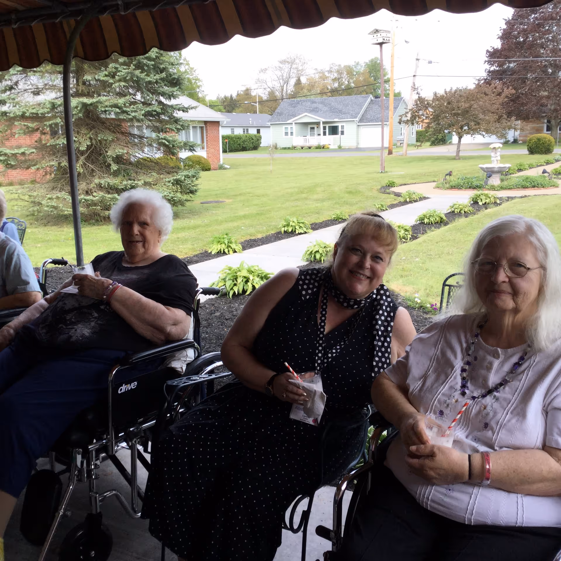 Three elderly women sitting outdoors under a canopy. Two of them are in wheelchairs, and all are holding drinks with striped straws. Behind them is a well-maintained lawn with a walkway, plants, trees, and houses in the background.