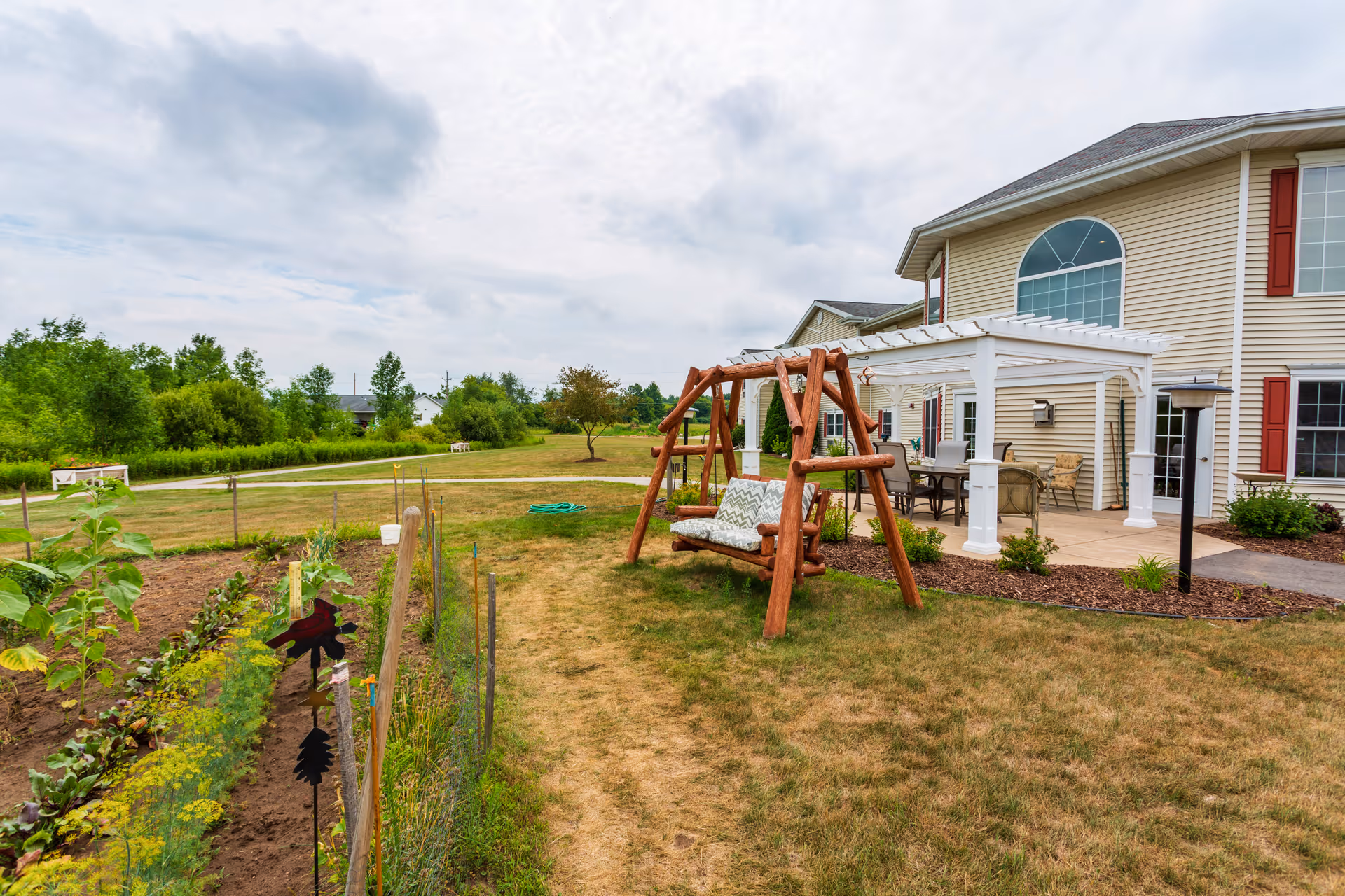 Outdoor garden area at The Cottages at Meadowlands Senior Living featuring a wooden swing with cushions, a pergola-covered patio with outdoor furniture, a vegetable garden with plants and stakes, and a large beige building with red shutters in the background under a cloudy sky.