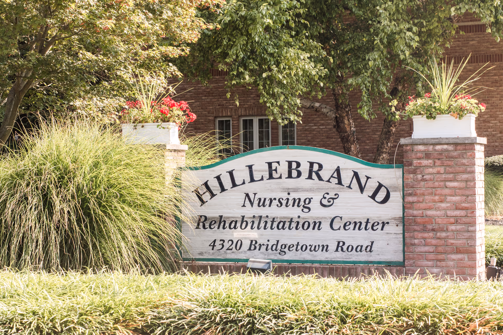 A wooden sign for Hillebrand Nursing & Rehabilitation Center at 4320 Bridgetown Road, surrounded by green plants and flowers, with a brick building and trees in the background.