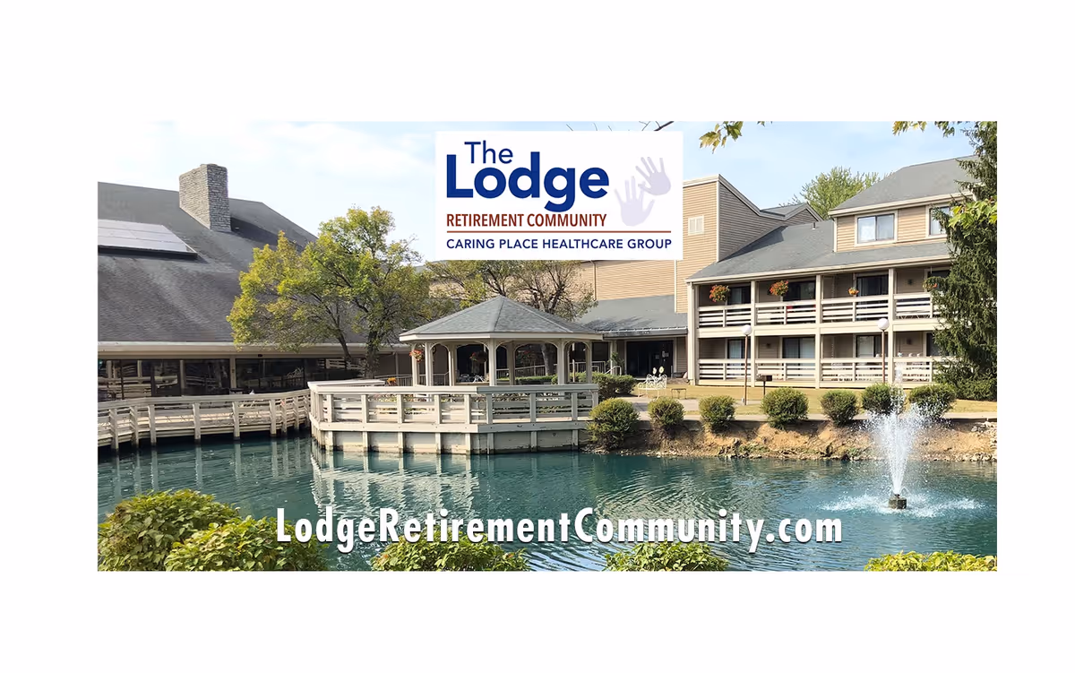 Exterior view of The Lodge Retirement Community featuring a pond with a fountain, a gazebo on a wooden deck, and multi-story buildings with balconies and flower pots.
