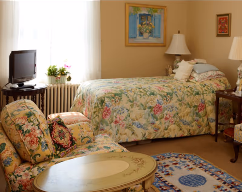 A cozy bedroom with a floral patterned bedspread and matching armchair. The room features a small round table, a TV on a wooden stand near a window with sheer white curtains, two table lamps, and framed artwork on the beige walls. A colorful rug is on the floor.