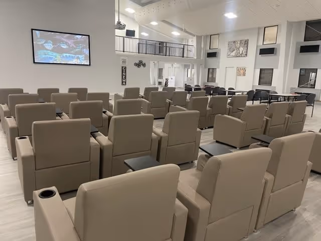 A spacious, modern theater room with multiple rows of beige reclining chairs facing a large screen mounted on the wall. The room has high ceilings with recessed lighting and a balcony area above. There are tables and chairs along the side walls, and decorative artwork is displayed on the walls.