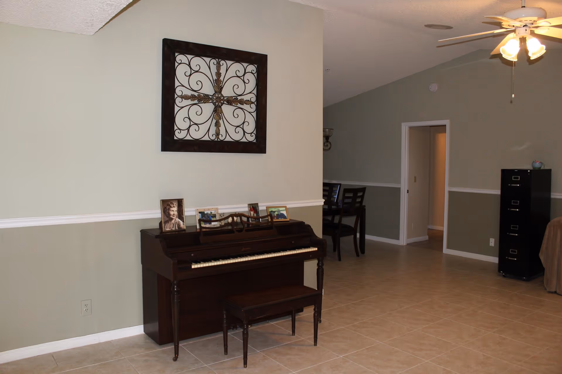 Interior view of a room in an assisted living home featuring a dark wooden piano with a matching bench. On top of the piano are framed photographs. Above the piano hangs a decorative wrought iron wall art. In the background, there is a dining area with chairs and a ceiling fan with lights. The walls are painted in two tones with a white chair rail, and the floor is tiled.