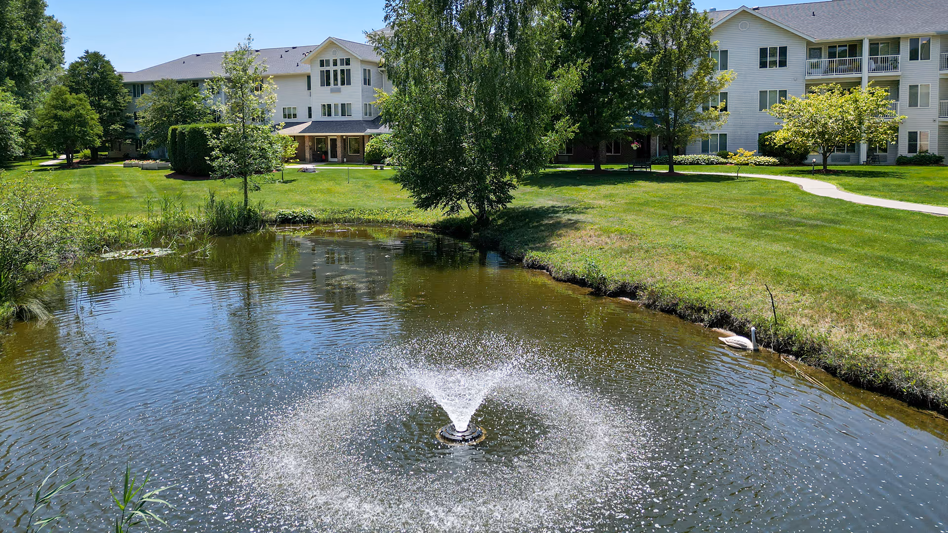 A serene outdoor scene at Holiday Blue Water Lodge featuring a pond with a central water fountain spraying water upwards. Surrounding the pond is a well-maintained grassy area with trees and shrubs. In the background, there are multi-story residential buildings with white siding and multiple windows under a clear blue sky.