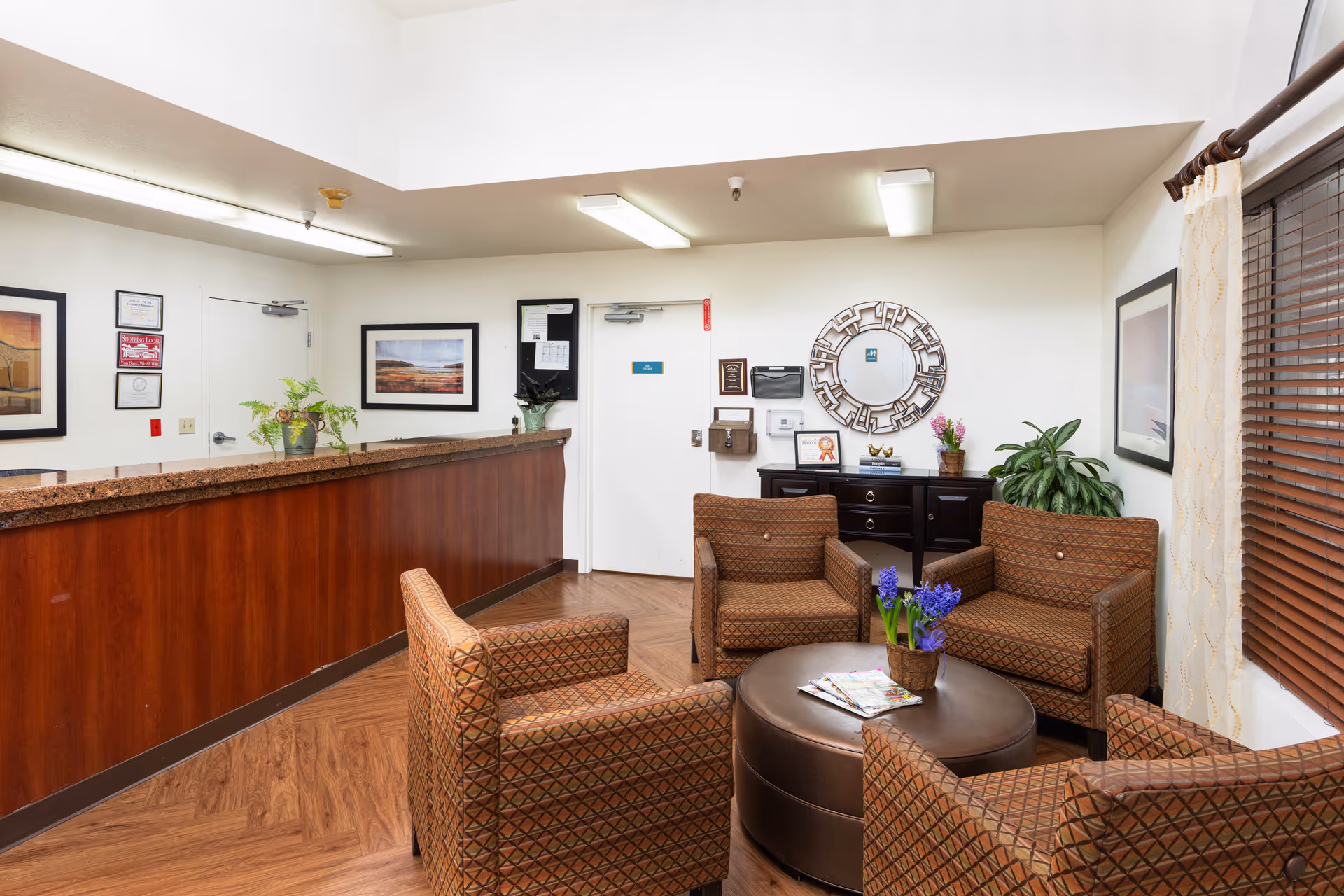 A reception area with a wooden front desk on the left and a seating area on the right featuring four patterned armchairs arranged around a round ottoman with a small plant and magazines on top. The walls are decorated with framed pictures, a round decorative mirror, and some certificates. There are plants on the desk and a cabinet against the wall. A window with wooden blinds and light curtains is visible on the right side.