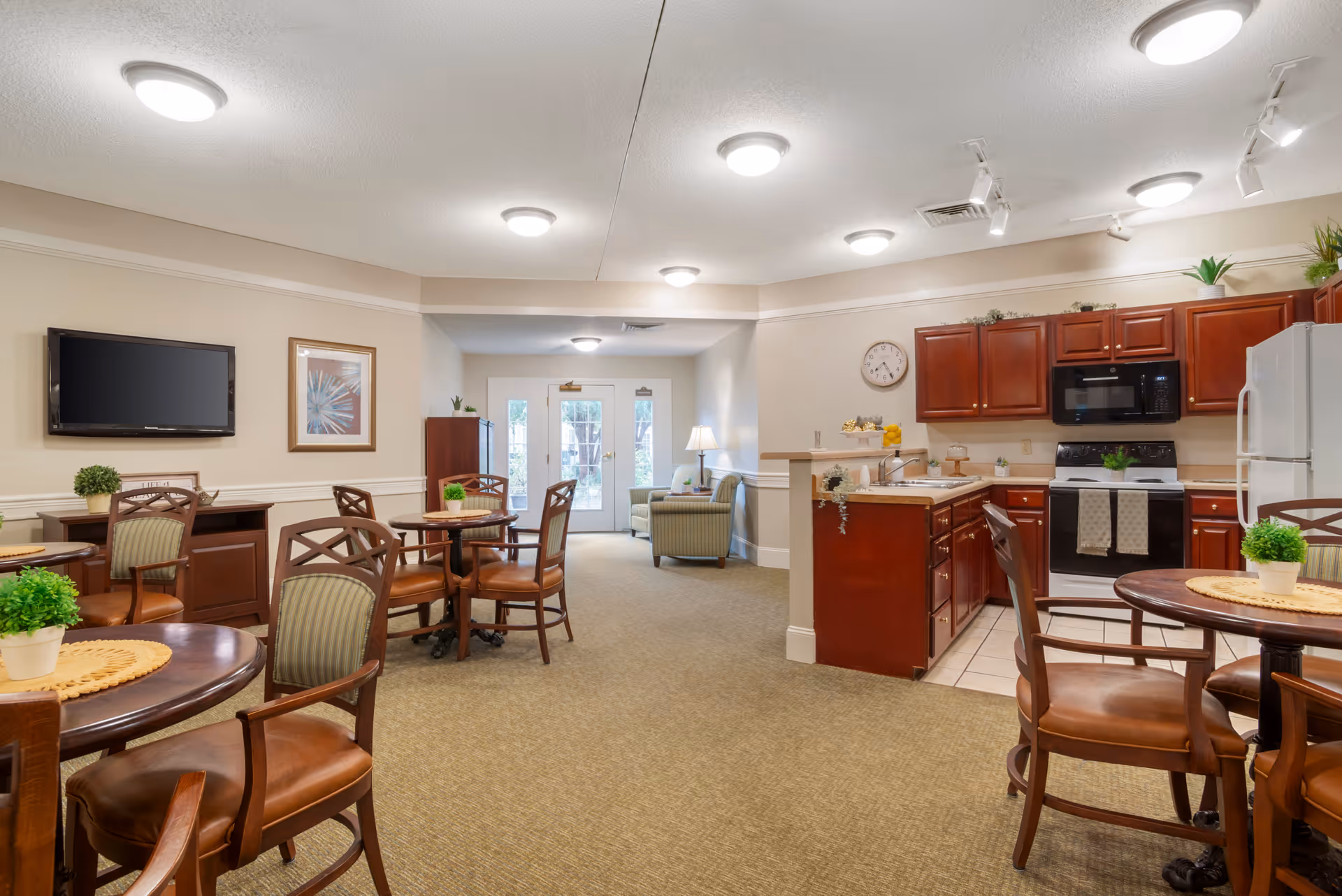 A bright and spacious common area in a senior living facility featuring a small kitchen with wooden cabinets, a stove, microwave, and refrigerator. Several round tables with chairs are arranged in the carpeted room, each table decorated with a small potted plant. A wall-mounted TV and framed artwork are visible on one wall. In the background, there is a seating area with armchairs near glass double doors leading outside.