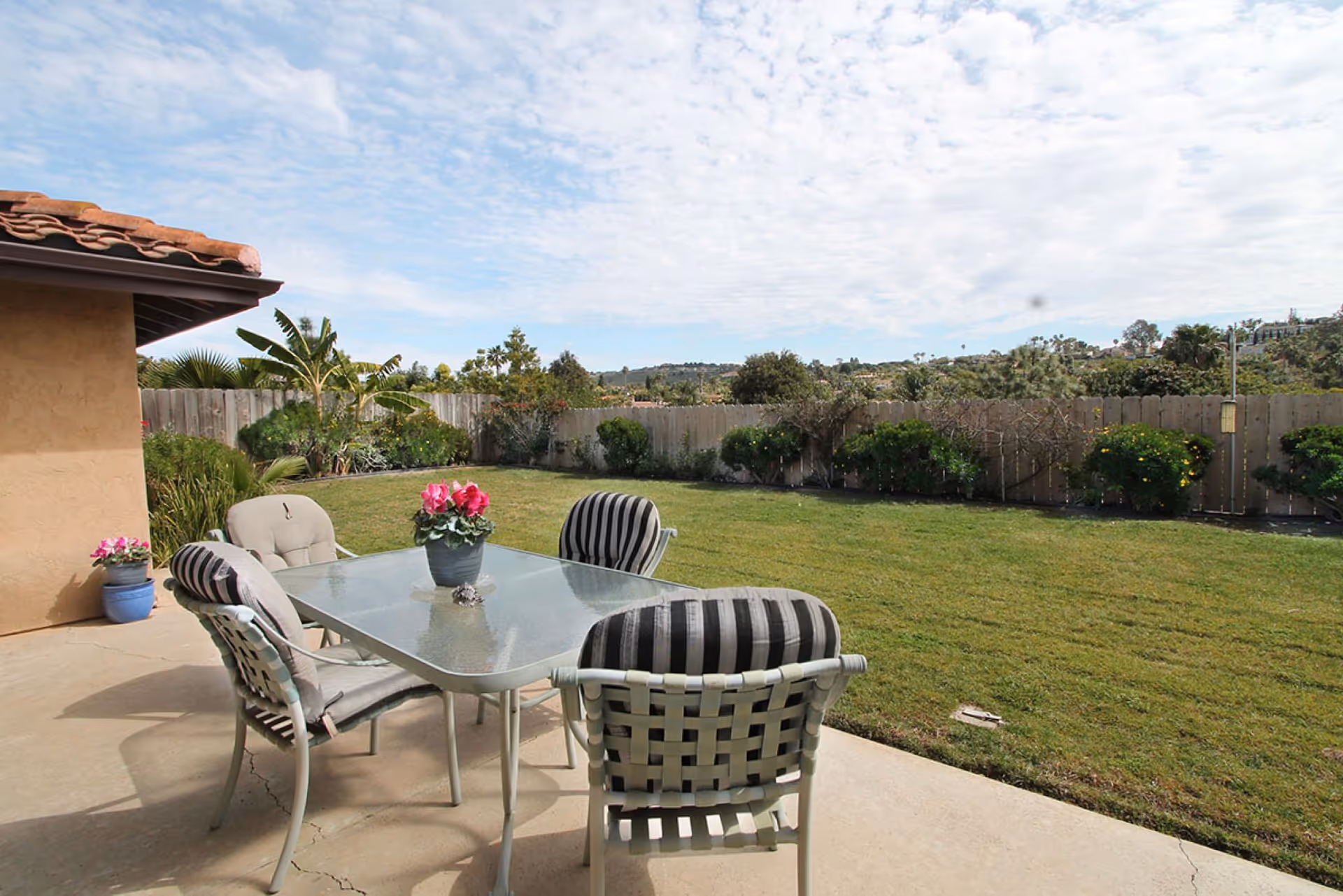 Patio with a glass table and four cushioned chairs overlooking a grassy fenced backyard with potted flowers and distant hills.