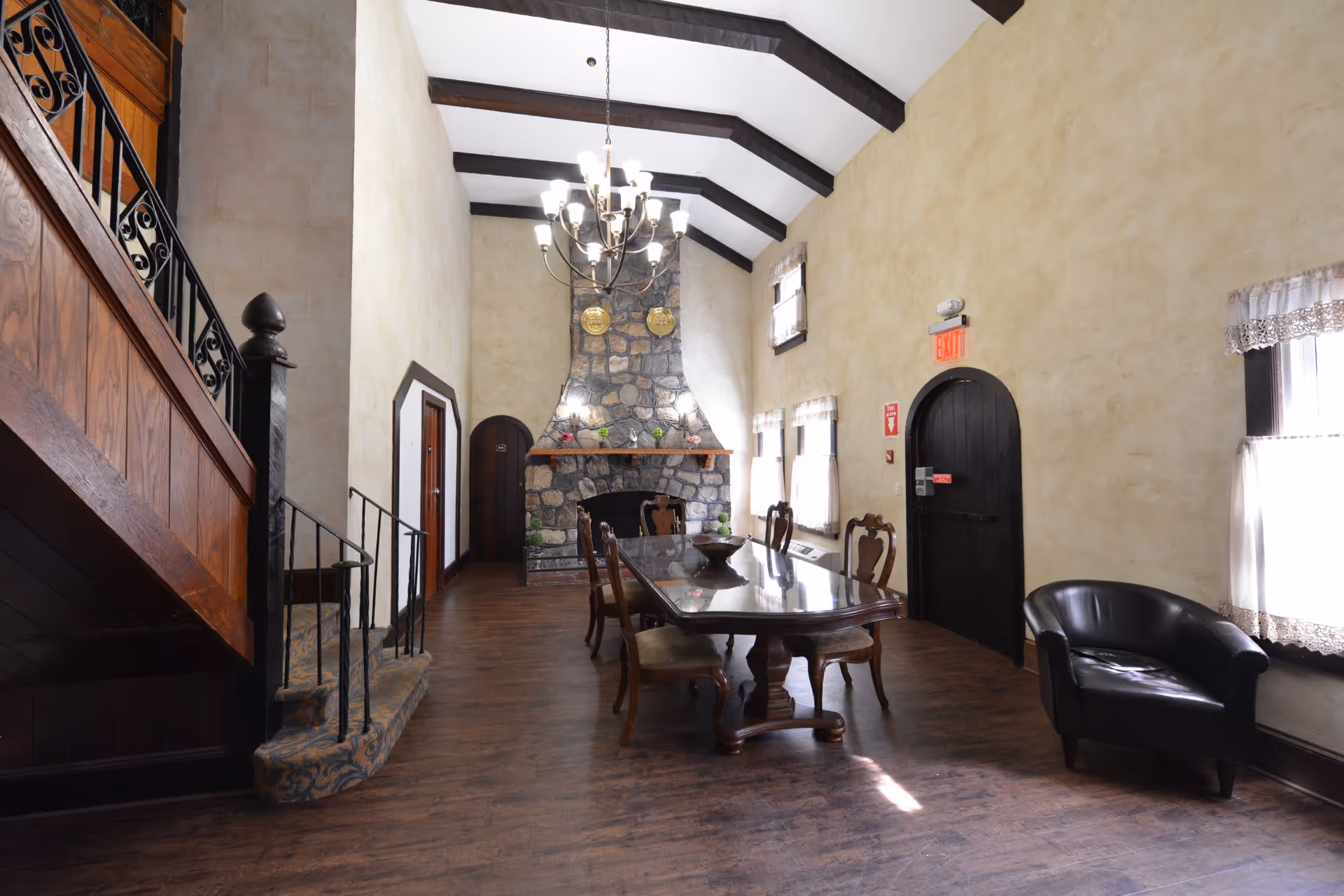 A spacious room with high vaulted ceilings featuring exposed dark wooden beams. There is a large stone fireplace at the far end with decorative plates above it. A long wooden dining table with six chairs is positioned in the center of the room. To the left, there is a wooden staircase with wrought iron railings. On the right side, there are three windows with lace curtains and a black leather armchair near the windows. The walls are painted a light beige color, and the floor is dark wood. An exit door with a red exit sign is visible on the right wall.
