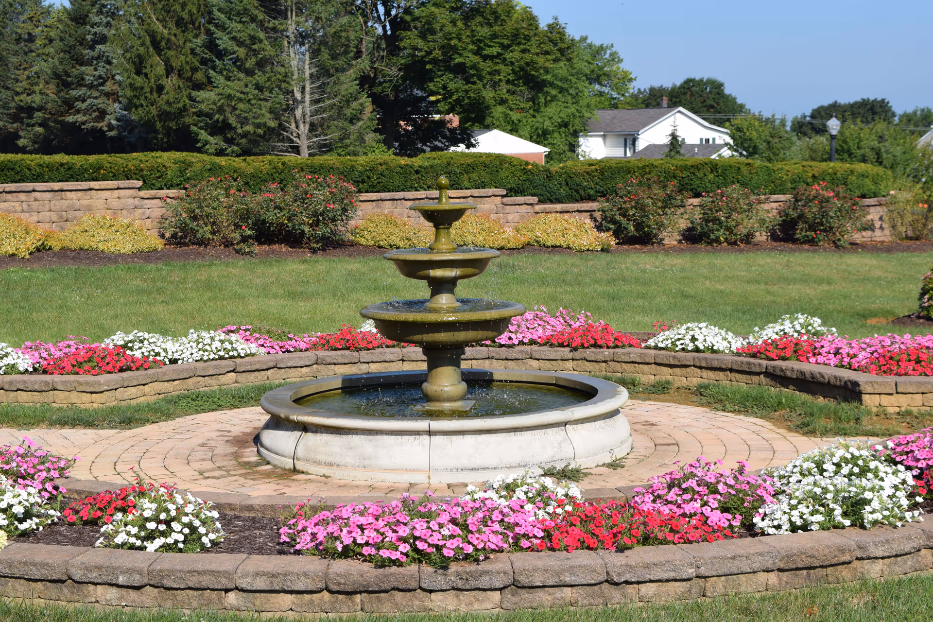 A three-tier stone fountain centered in a circular paved area surrounded by colorful flower beds and manicured lawn.