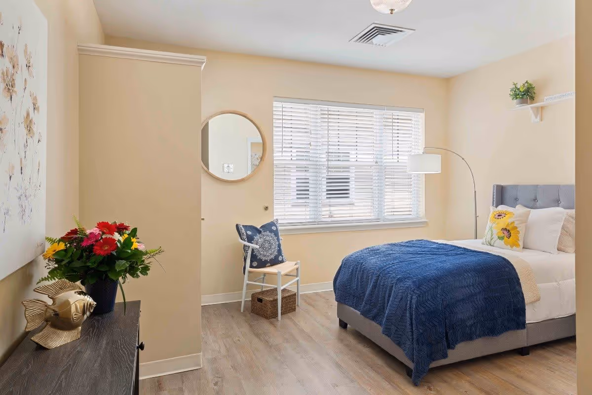 A bright and cozy bedroom with a bed covered in a blue blanket and white pillows, a chair with a decorative pillow, a round wall mirror, a floor lamp, a small shelf with a plant, and a dresser with a vase of colorful flowers and a gold fish sculpture. The room has light-colored walls and wood flooring.