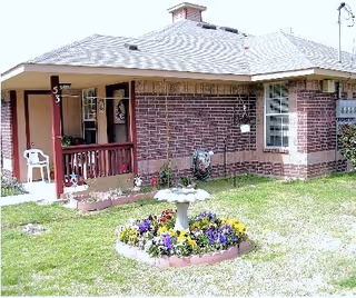 A single-story brick senior living facility with a small front porch, a white plastic chair, and a circular flower bed in the lawn.