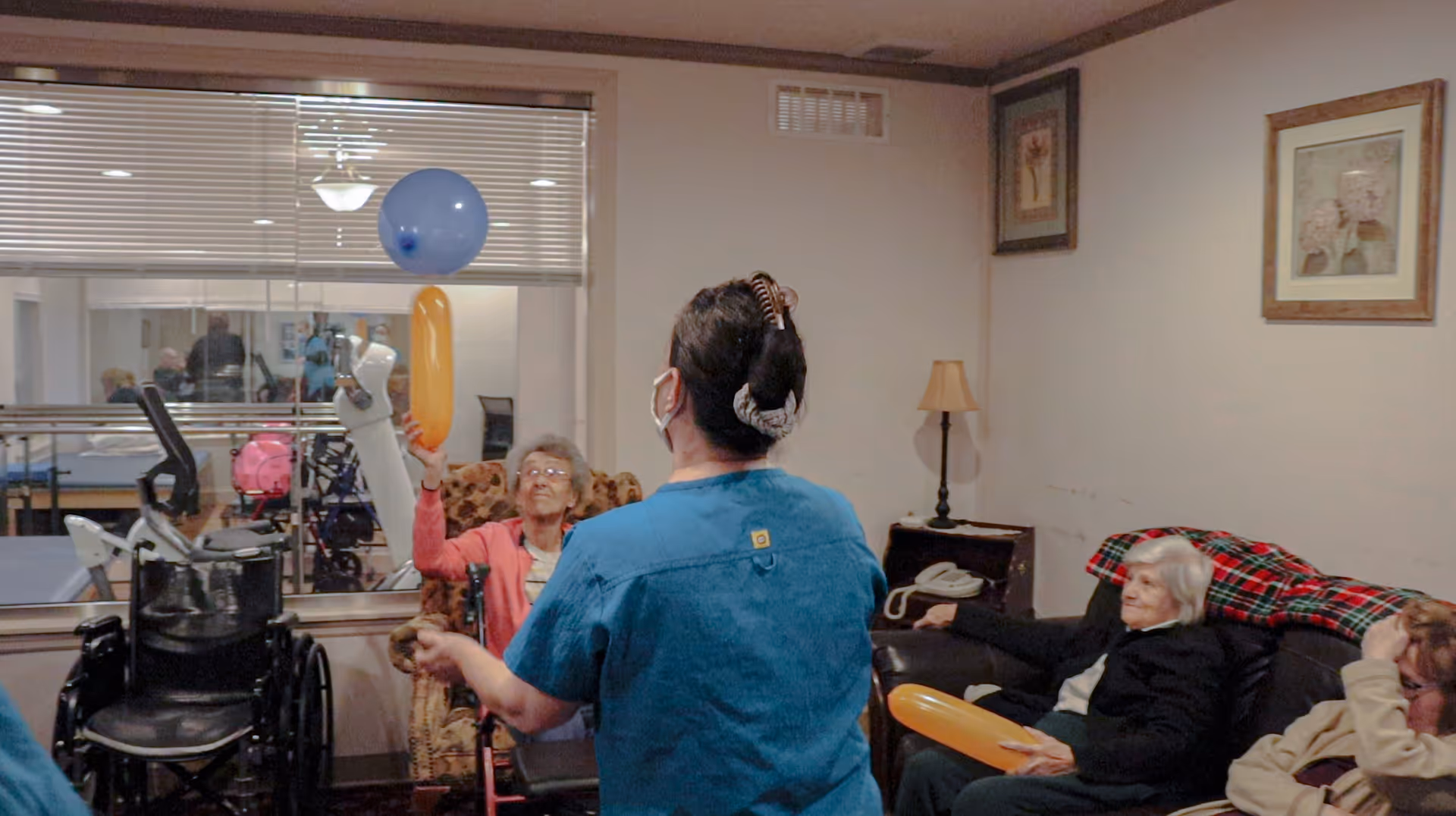 A group of elderly people and a caregiver in a common room of a senior living facility. The caregiver, wearing a blue uniform and a face mask, is engaging with the residents by tossing balloons. Some elderly individuals are seated on chairs and a couch, participating in the activity. The room has framed pictures on the walls, a lamp on a side table, and a window showing exercise equipment in another room.