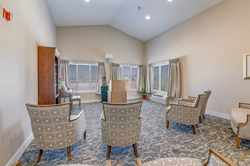 A bright and spacious common area in a senior living facility with patterned armchairs arranged in a semi-circle facing a wooden podium. The room features large windows with curtains, a carpeted floor with a blue and beige pattern, and light-colored walls. There is a wooden bookshelf and a piano against one wall, and a small green cabinet with a plant on top near the windows.