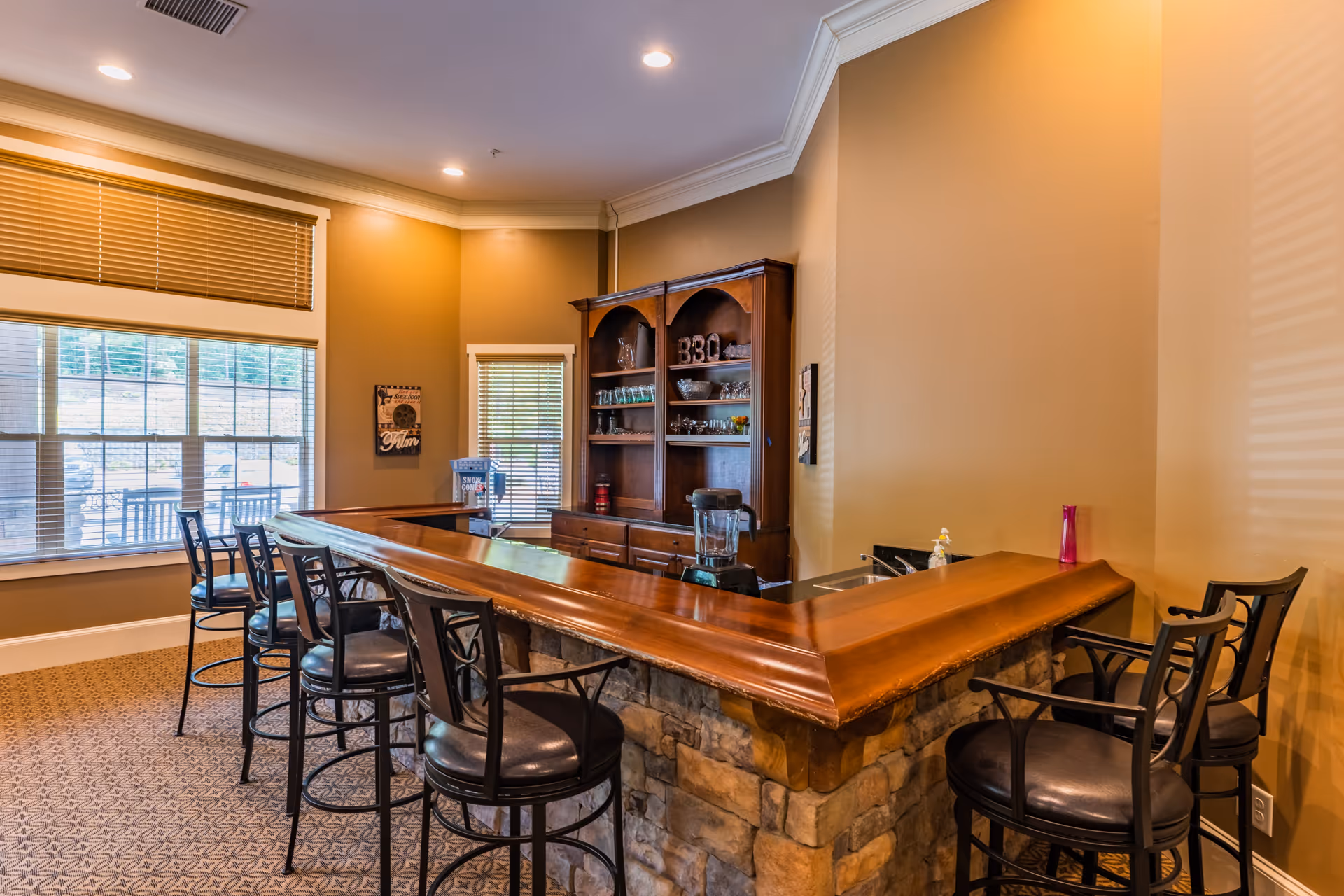 Interior view of a senior living facility's bar area with a wooden counter, stone base, and several black bar stools. Behind the counter is a wooden cabinet with glassware and decorative items. Large windows with blinds allow natural light into the room, which has beige walls and patterned carpet flooring.