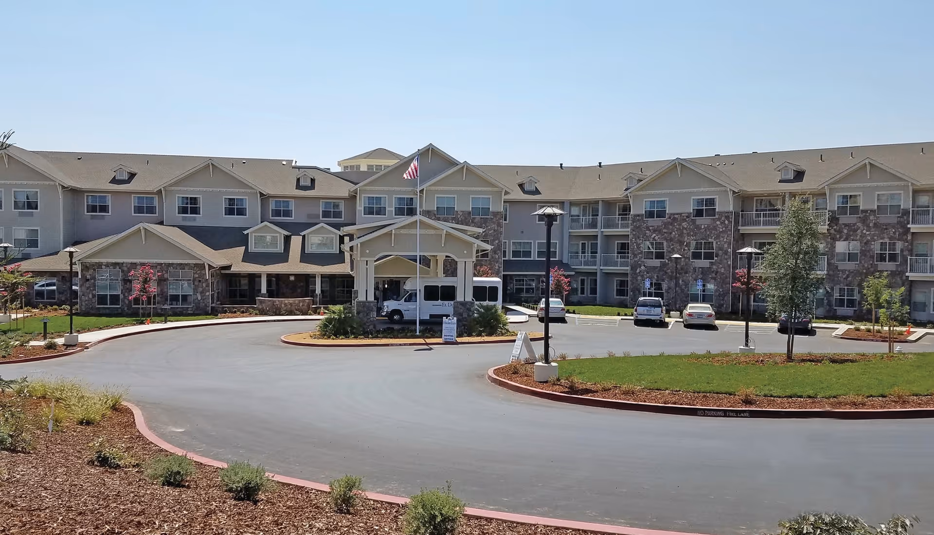 Front exterior view of El Dorado Estates Gracious Retirement Living facility with a circular driveway, landscaped greenery, and several parked vehicles under a clear blue sky.