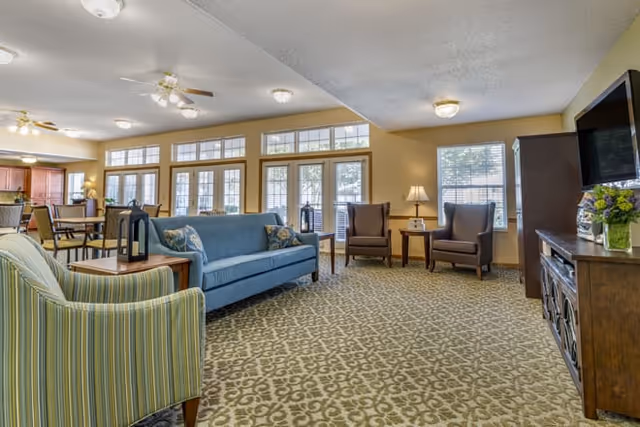 A spacious and well-lit living room area in an assisted living facility featuring a blue sofa, two brown armchairs, a striped armchair, a wooden TV stand with a flat-screen TV, and large windows allowing natural light to fill the room. The room has patterned carpet flooring and ceiling fans with lights.