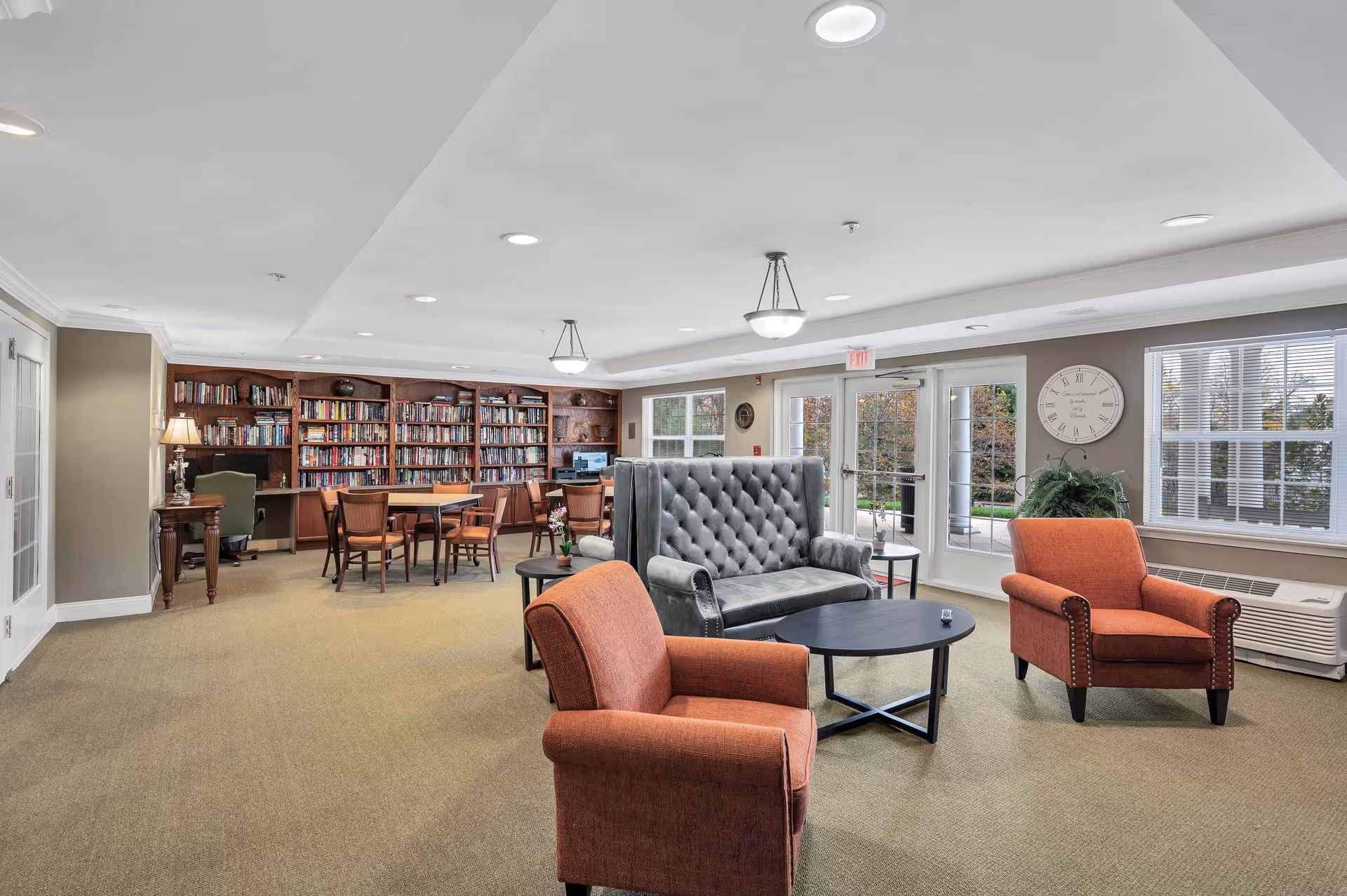 A spacious and well-lit common area with a seating arrangement including two orange armchairs and a gray tufted loveseat around a black coffee table. In the background, there is a large wooden bookshelf filled with books, a table with chairs, and a computer workstation. Large windows and glass doors let in natural light and offer a view of the outdoor greenery.