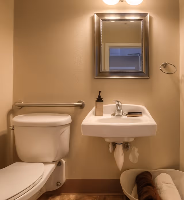 A small bathroom with a white toilet and a wall-mounted white sink. Above the sink is a rectangular mirror with a silver frame and a light fixture above it. There is a soap dispenser and a bar of soap on the sink. A towel ring is mounted on the wall to the right of the sink, and a grab bar is installed on the wall behind the toilet. A basket with rolled towels is placed on the floor to the right of the sink.
