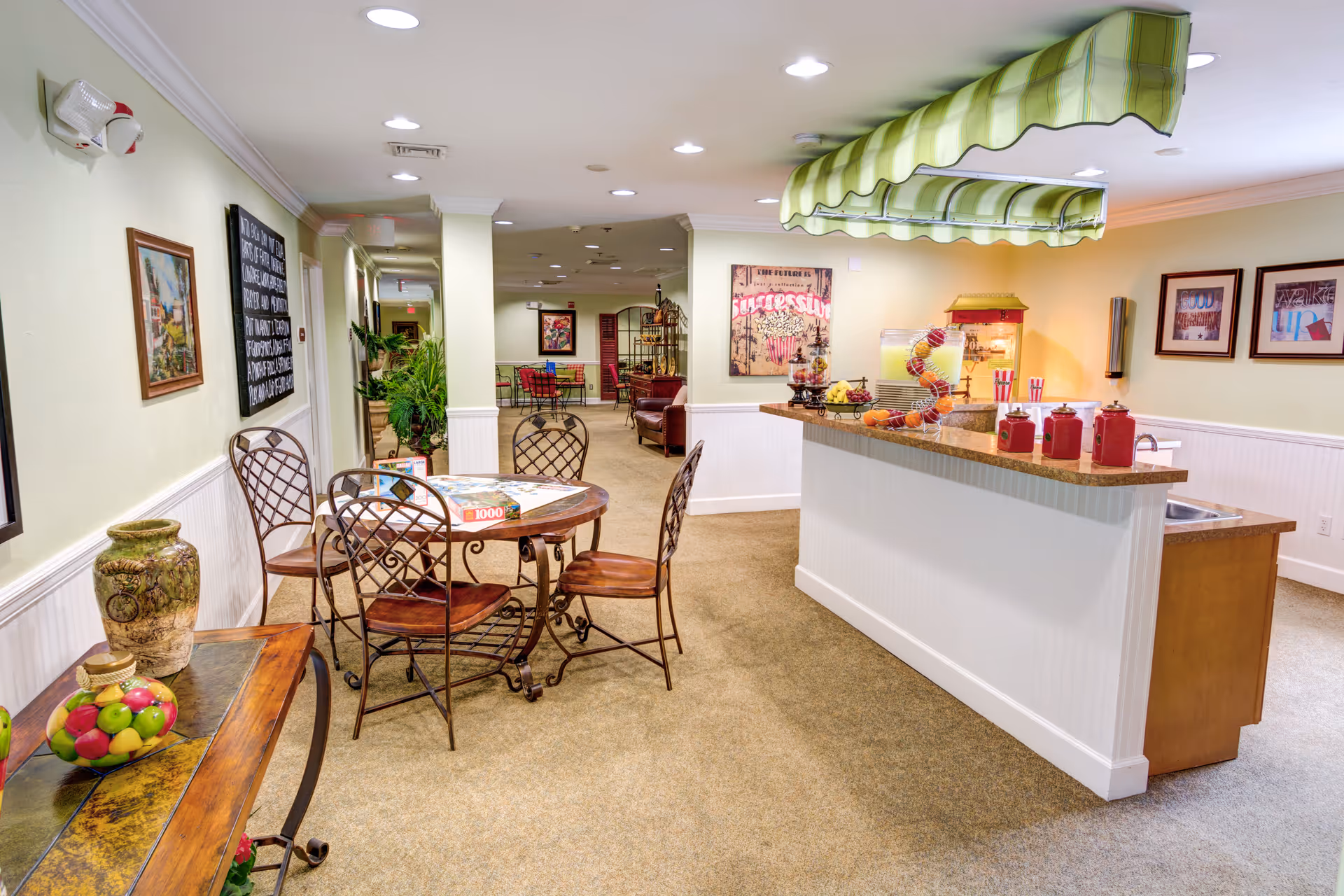 Bright communal dining and snack area with a round table and chairs, a service counter with a green striped canopy, and wall artwork.