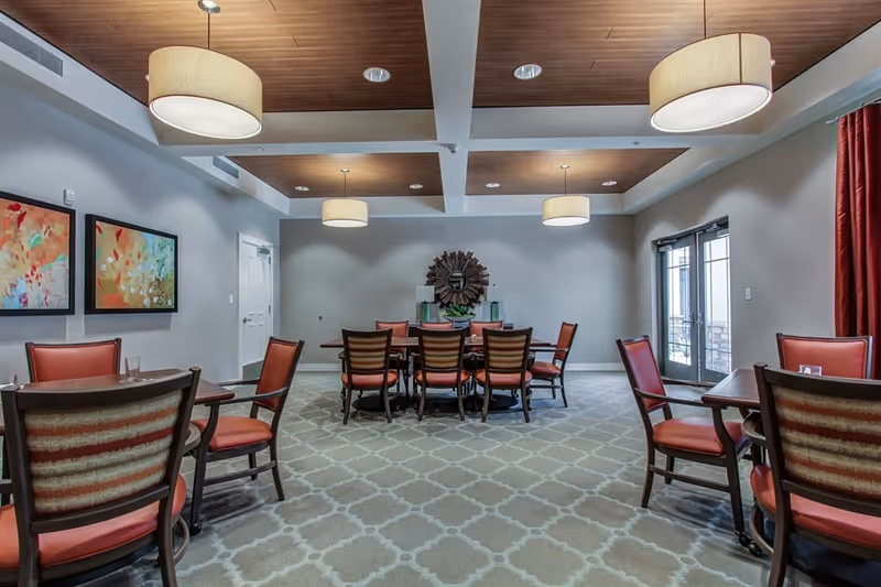 A dining room with multiple wooden tables and chairs with red cushions. The room has a patterned carpet, a wooden ceiling with recessed lighting and hanging lamps, two colorful abstract paintings on the left wall, and a decorative mirror on the far wall. There are glass doors on the right side with red curtains.
