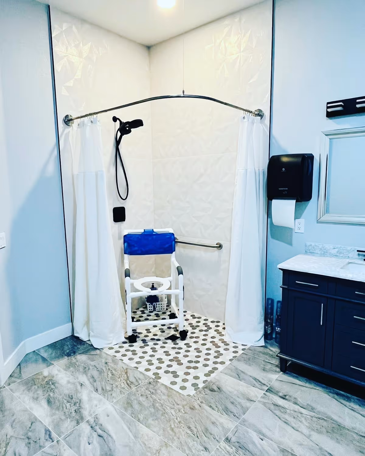 A bathroom with a walk-in shower featuring a white and blue shower chair, a curved shower curtain rod with white curtains, and a handheld showerhead. The shower area has white textured wall tiles and hexagonal floor tiles. To the right, there is a dark-colored vanity with a marble countertop, a mounted paper towel dispenser, and a mirror.