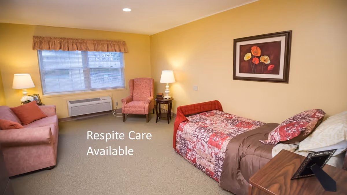 A cozy bedroom in a senior living facility with a single bed covered in a patterned quilt, a pink armchair, a pink sofa with cushions, two table lamps, a window with blinds and a valance, and a framed floral painting on the wall. Text on the image reads 'Respite Care Available'.