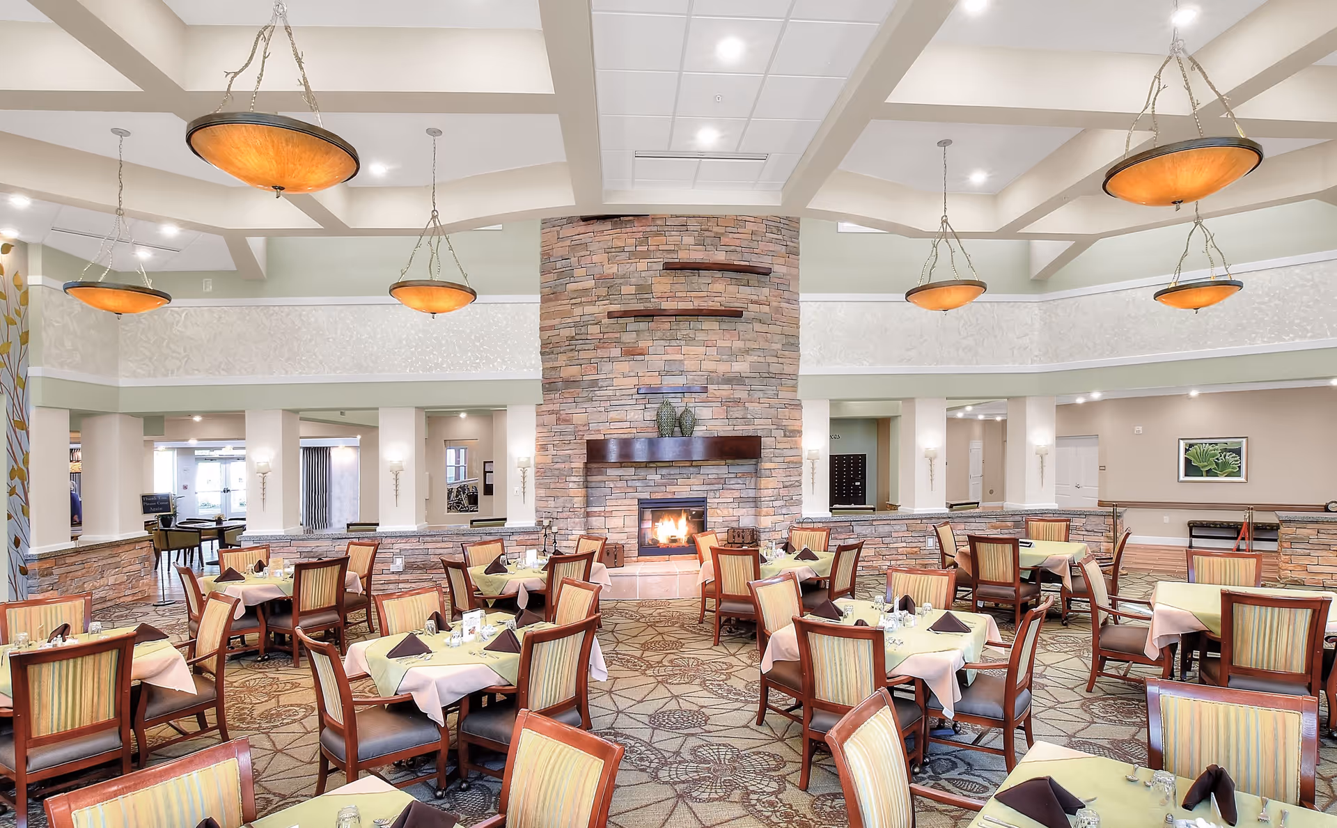 A spacious dining room in a senior living facility with multiple tables covered in light green and beige tablecloths, each set with napkins and glassware. The room features a large stone fireplace in the center with decorative shelves above it. The ceiling has exposed beams and several hanging pendant lights with warm amber shades. The walls are painted in soft neutral tones, and the floor is carpeted with a patterned design.