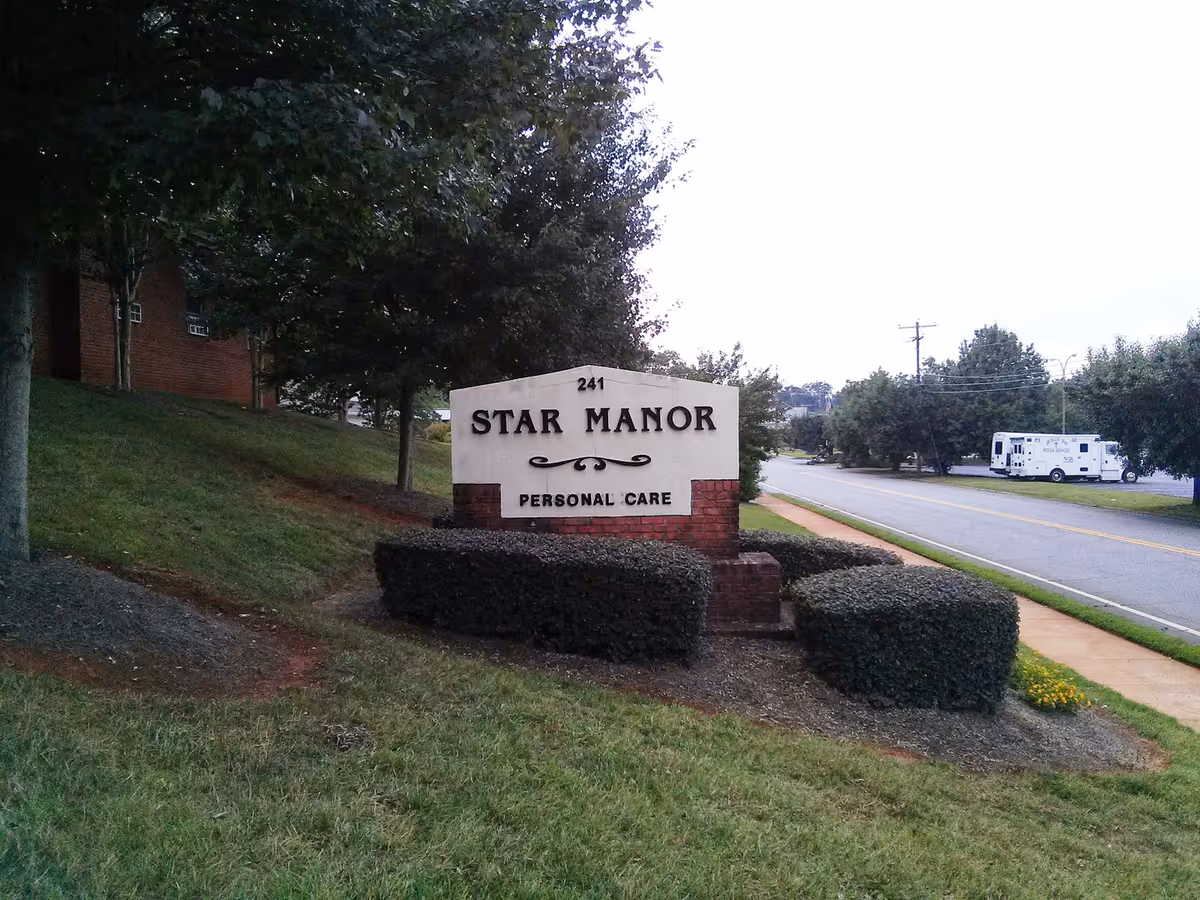 Outdoor view of a sign for Star Manor Personal Care located on a grassy area with trimmed bushes around it. There are trees and a sidewalk next to a road with a parked emergency vehicle in the background.