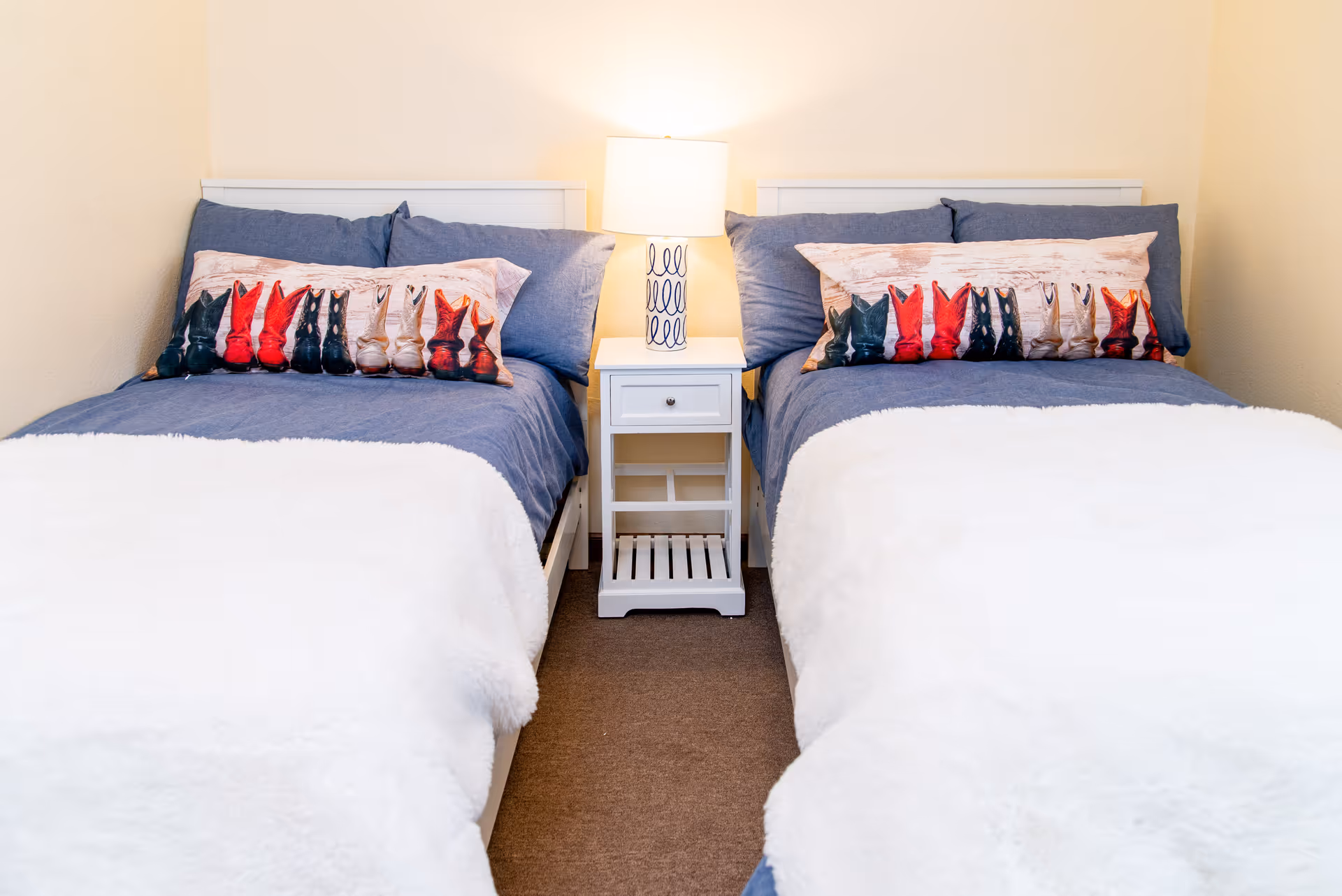 Two twin beds with blue bedding and white fluffy blankets, each adorned with a decorative pillow featuring a pattern of colorful cowboy boots. A white nightstand with a drawer and shelf sits between the beds, topped with a white lamp with a blue swirl design. The room has beige walls and a brown carpeted floor.