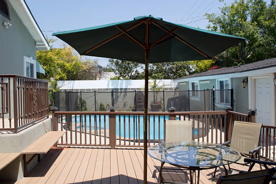 Outdoor patio area with a round glass table, four chairs, and a large green umbrella providing shade. The patio overlooks a fenced swimming pool surrounded by a white privacy fence and greenery.