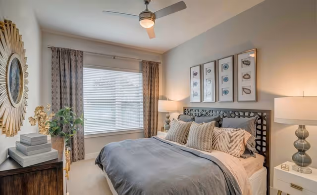 A cozy bedroom with a large bed featuring multiple pillows and a gray comforter. There is a window with patterned curtains on the left wall, a decorative round mirror above a wooden dresser with stacked books and a plant, and two bedside tables each with a lamp. Above the bed are four framed artworks with shell designs. The room has a ceiling fan with a light.