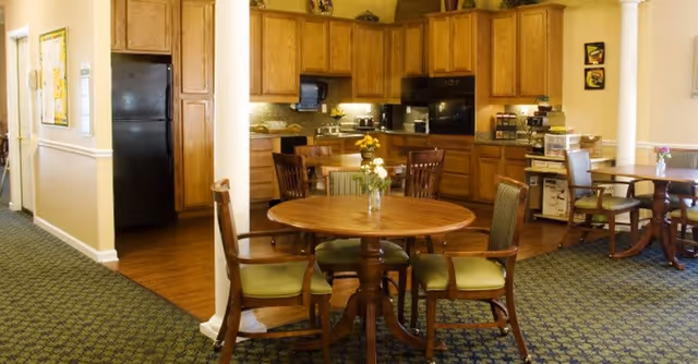 A cozy kitchen and dining area in a senior living facility with wooden cabinets, a black refrigerator, and a round wooden table surrounded by four chairs with green cushions. The room has patterned carpet flooring and soft yellow walls, with small flower vases on the tables.