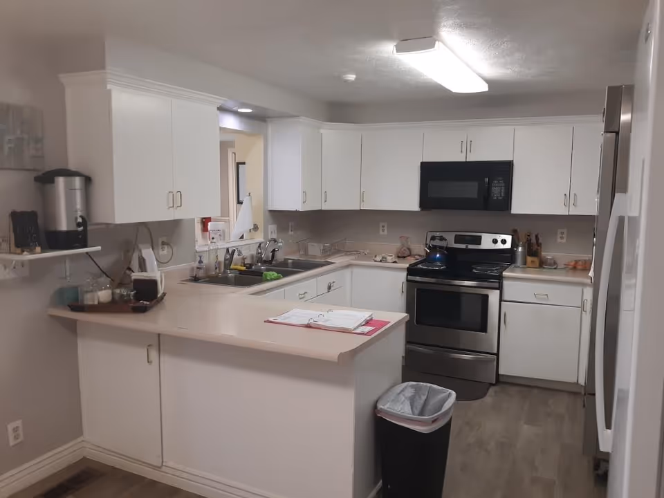 A clean and modern kitchen with white cabinets, a stainless steel oven and microwave, a double sink, and a countertop with a binder and some kitchen items. There is a trash can on the floor and a water dispenser on a small wall shelf to the left.