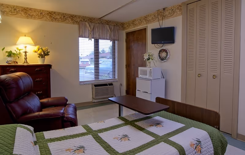 A nursing facility bedroom with a bed covered in a floral quilt, an overbed table, a leather recliner, a dresser with lamp and plants, a small microwave on a cabinet, a wall-mounted TV, window and closet doors.