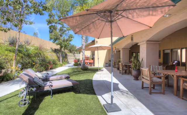 Outdoor patio area at a senior living facility with lounge chairs, umbrellas, tables, and chairs on a concrete and artificial grass surface, surrounded by plants and trees under a clear blue sky.