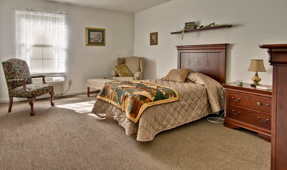 Sunlit bedroom with a single bed dressed in a quilt, wooden headboard and nightstand, two armchairs and a window air conditioner.