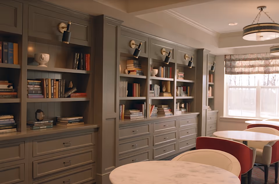 Cozy common room with built-in bookshelves, round marble-top tables and upholstered chairs by a window.