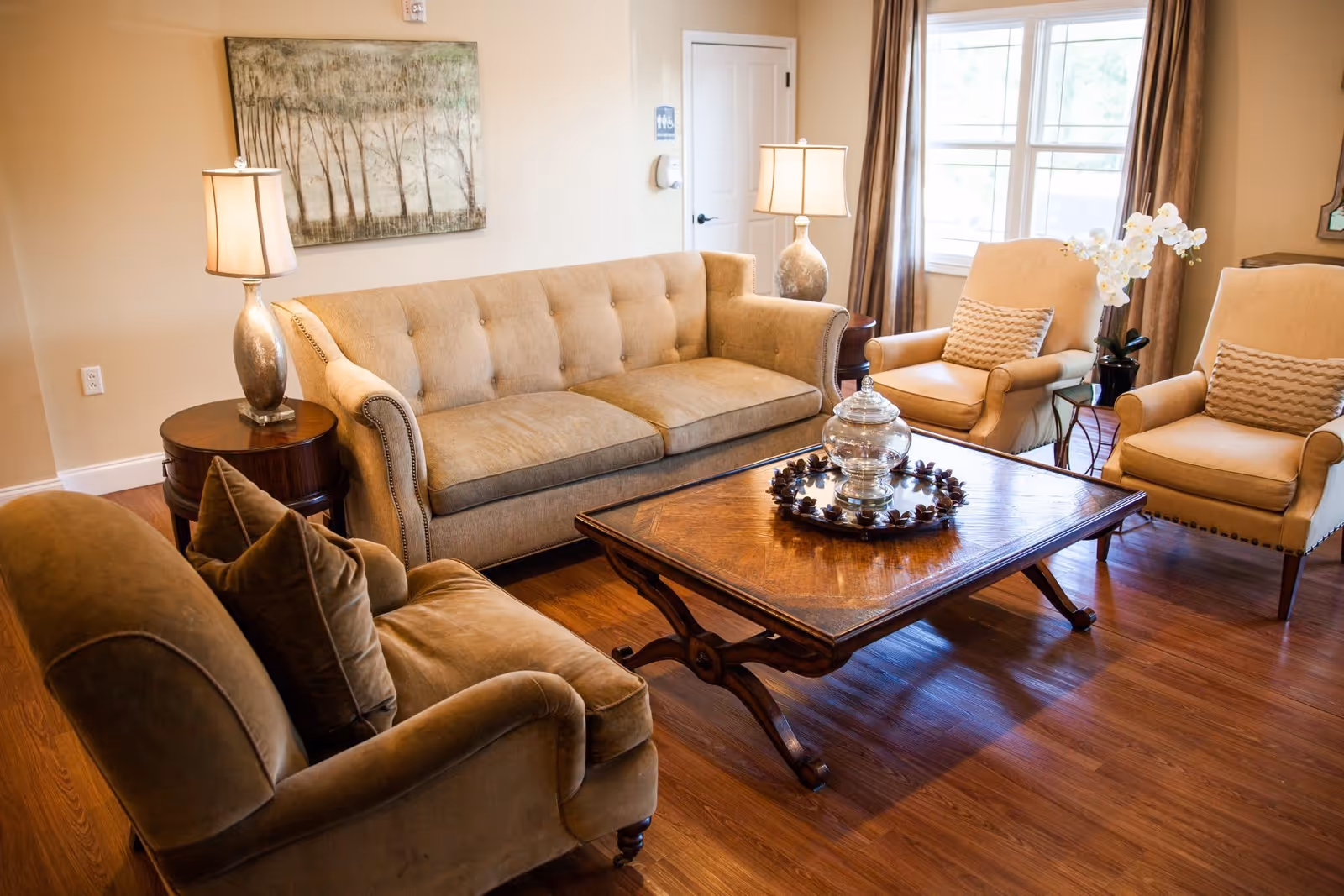A cozy living room with a beige sofa, two beige armchairs with patterned cushions, and a brown armchair with matching pillows. A wooden coffee table with a decorative glass jar and a circular arrangement of pinecones sits in the center. Two table lamps with beige shades are on round wooden side tables. A large window with brown curtains lets in natural light, and a painting of trees hangs on the wall.