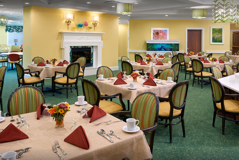 Bright assisted-living dining room with multiple round tables set with beige tablecloths, folded red napkins, floral centerpieces, and striped chairs.