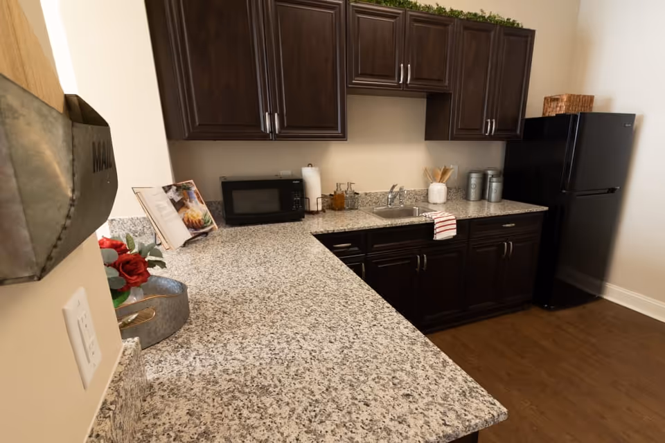 A kitchen area with dark wood cabinets, a granite countertop, a black microwave, a small sink with a striped towel hanging on the cabinet handle, a black refrigerator, and various kitchen items including a cookbook, paper towel holder, and containers. The floor is wooden and there is a metal mail holder and a small flower arrangement on the left side.