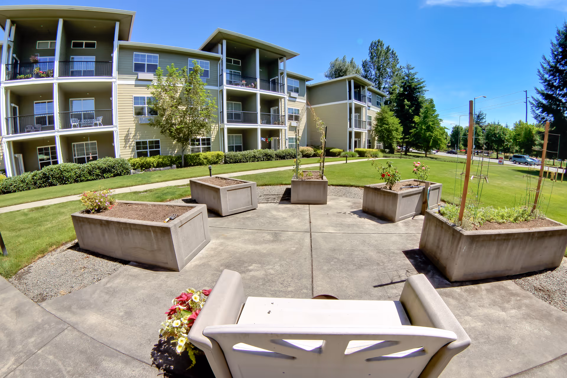Outdoor garden area with several rectangular concrete planters containing small plants and flowers, surrounded by a paved circular patio. In the foreground, there is a white bench with flowers beside it. In the background, there is a three-story residential building with balconies, green grass, trees, and a clear blue sky.