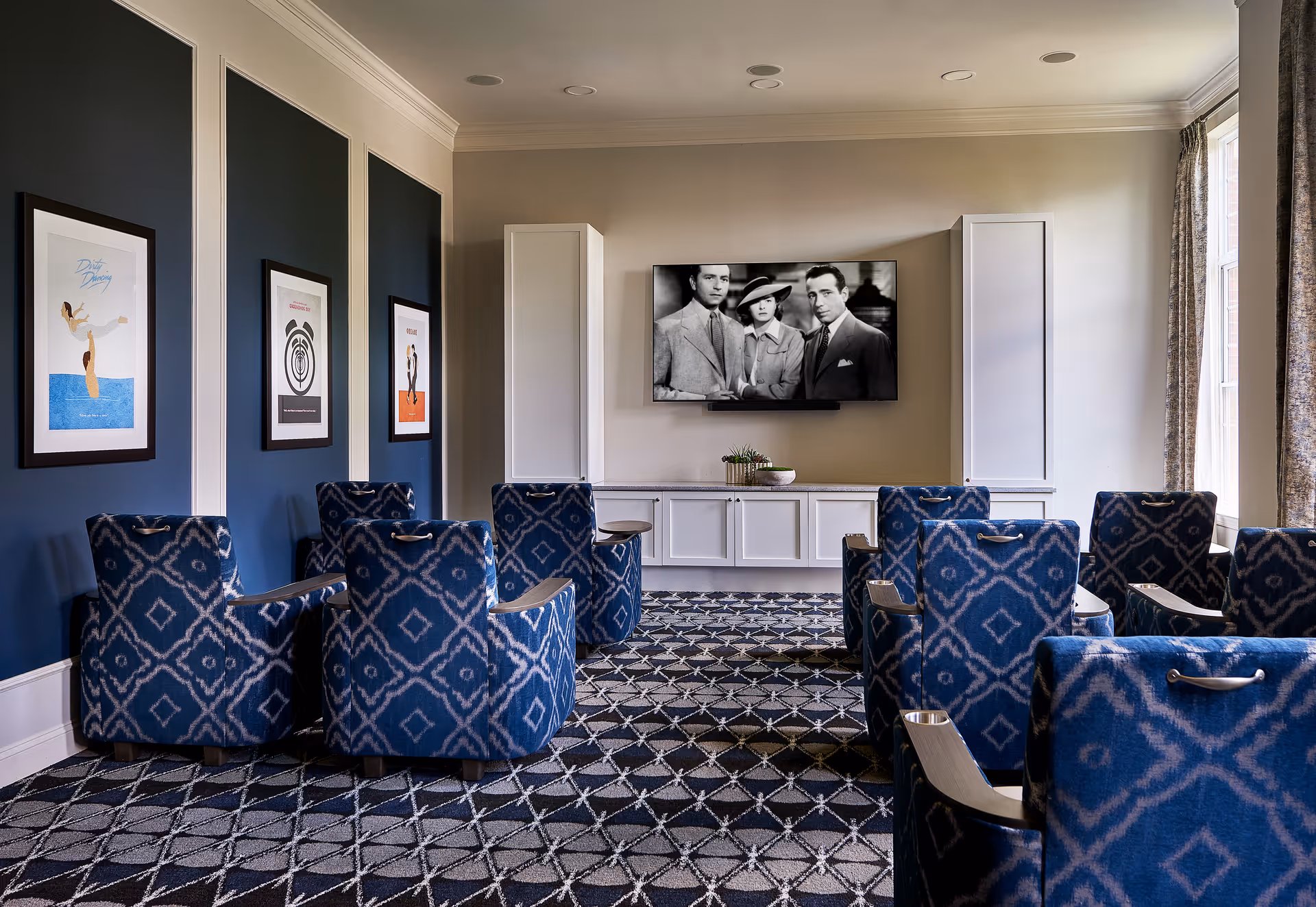 A cozy media room with patterned blue armchairs arranged in rows facing a wall-mounted flat-screen TV displaying a black and white movie scene. The room features dark blue accent walls with framed artwork, white cabinetry below the TV, and large windows with curtains allowing natural light to enter.
