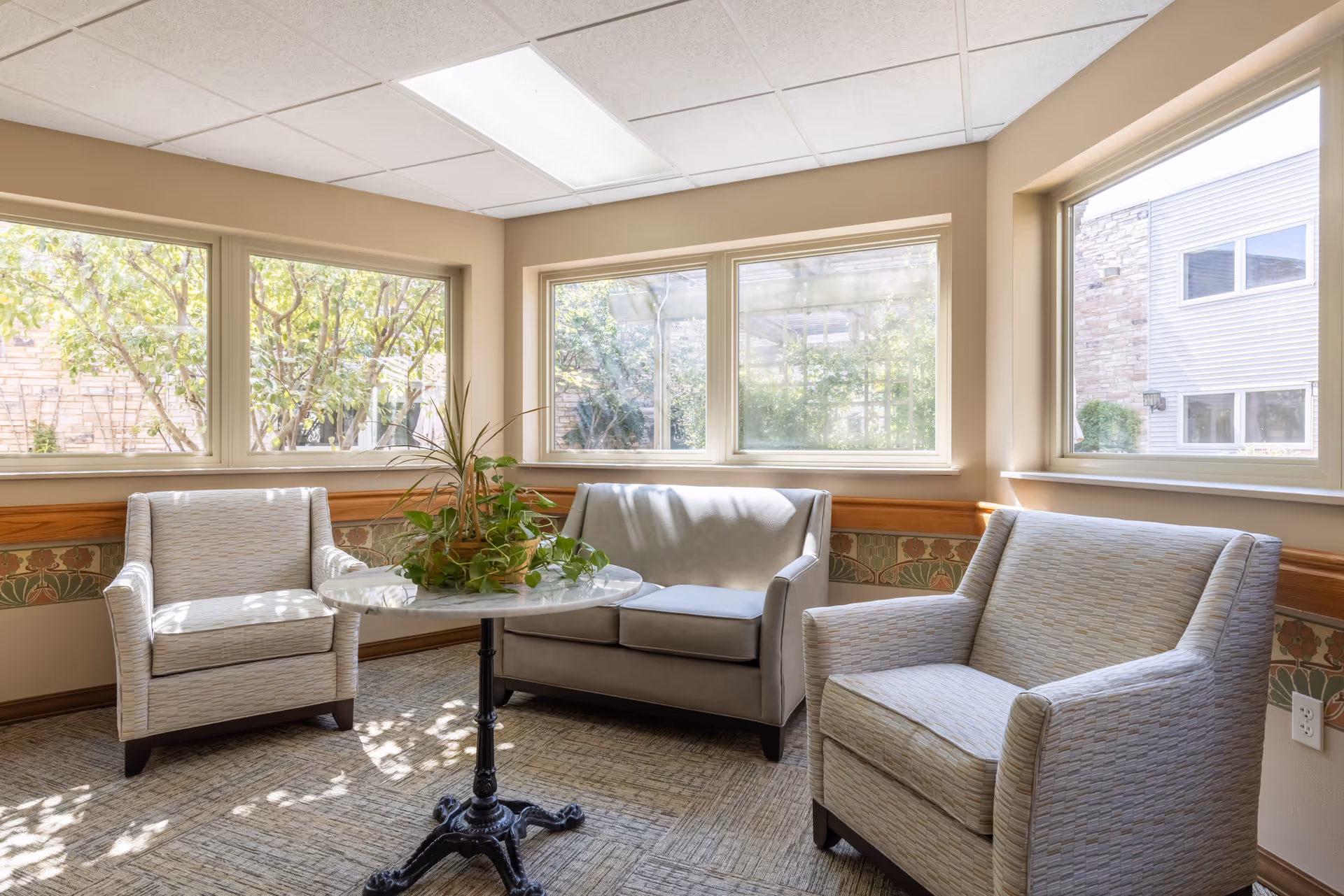 A bright sitting area with three upholstered chairs arranged around a small round table with a plant centerpiece. The room has large windows letting in natural light and a view of greenery outside.