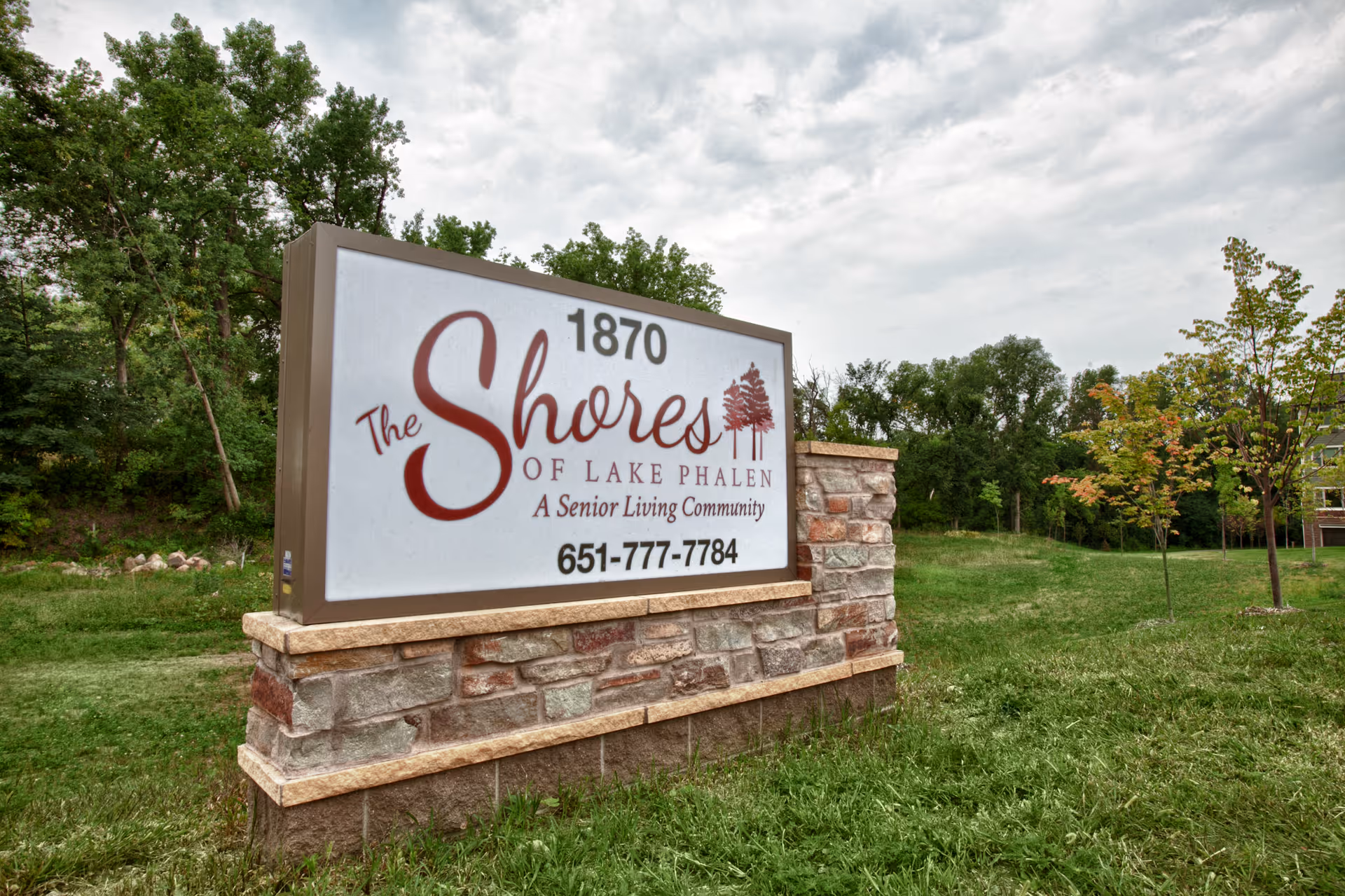 A large outdoor sign for The Shores of Lake Phalen, a senior living community, set on a stone base with grass and trees in the background under a cloudy sky.