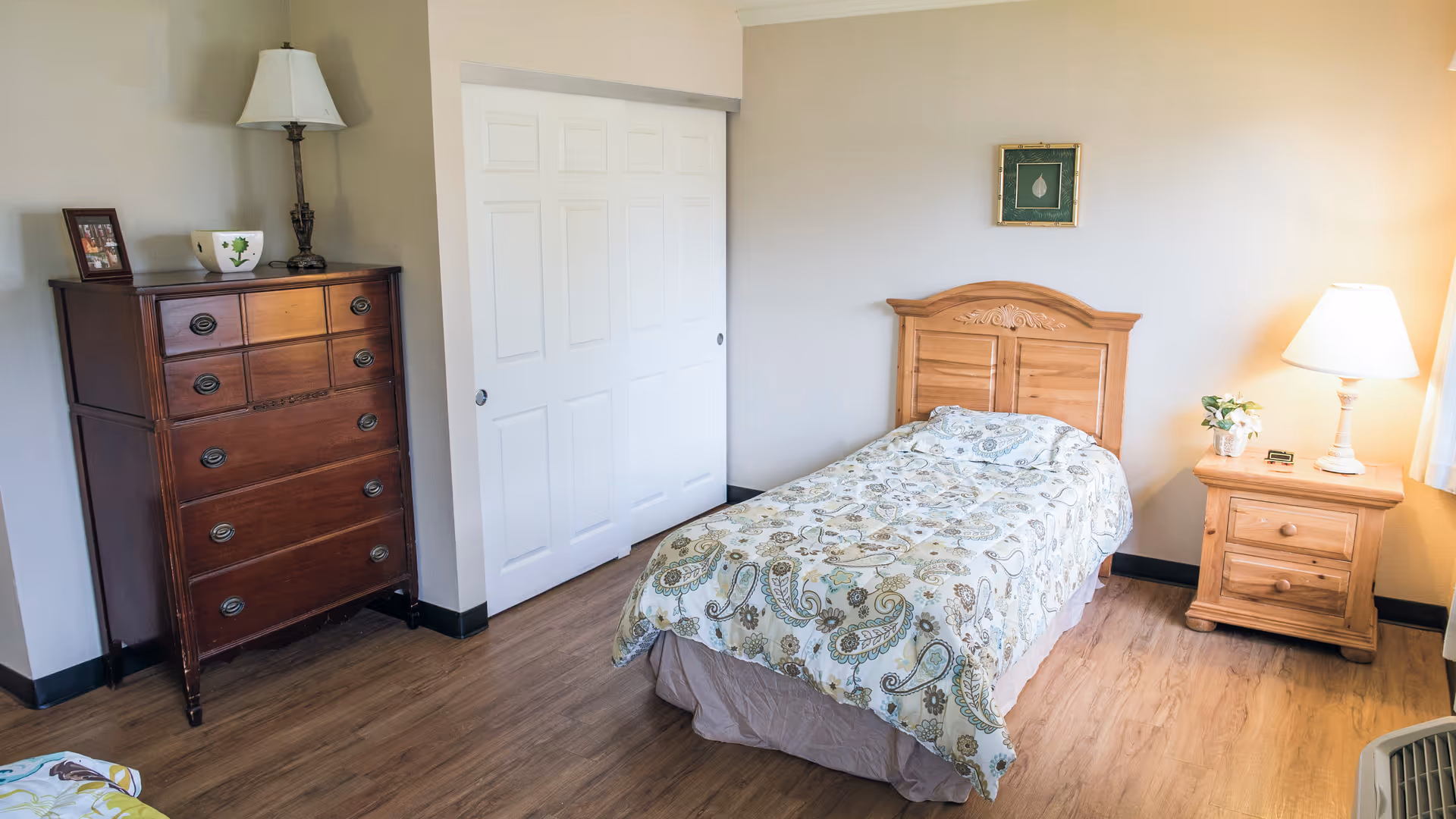 A simple bedroom with a single bed covered with a patterned comforter, a wooden headboard, a wooden nightstand with a lamp and a small plant, a wooden dresser with a lamp and a framed photo, and a closet with white sliding doors.