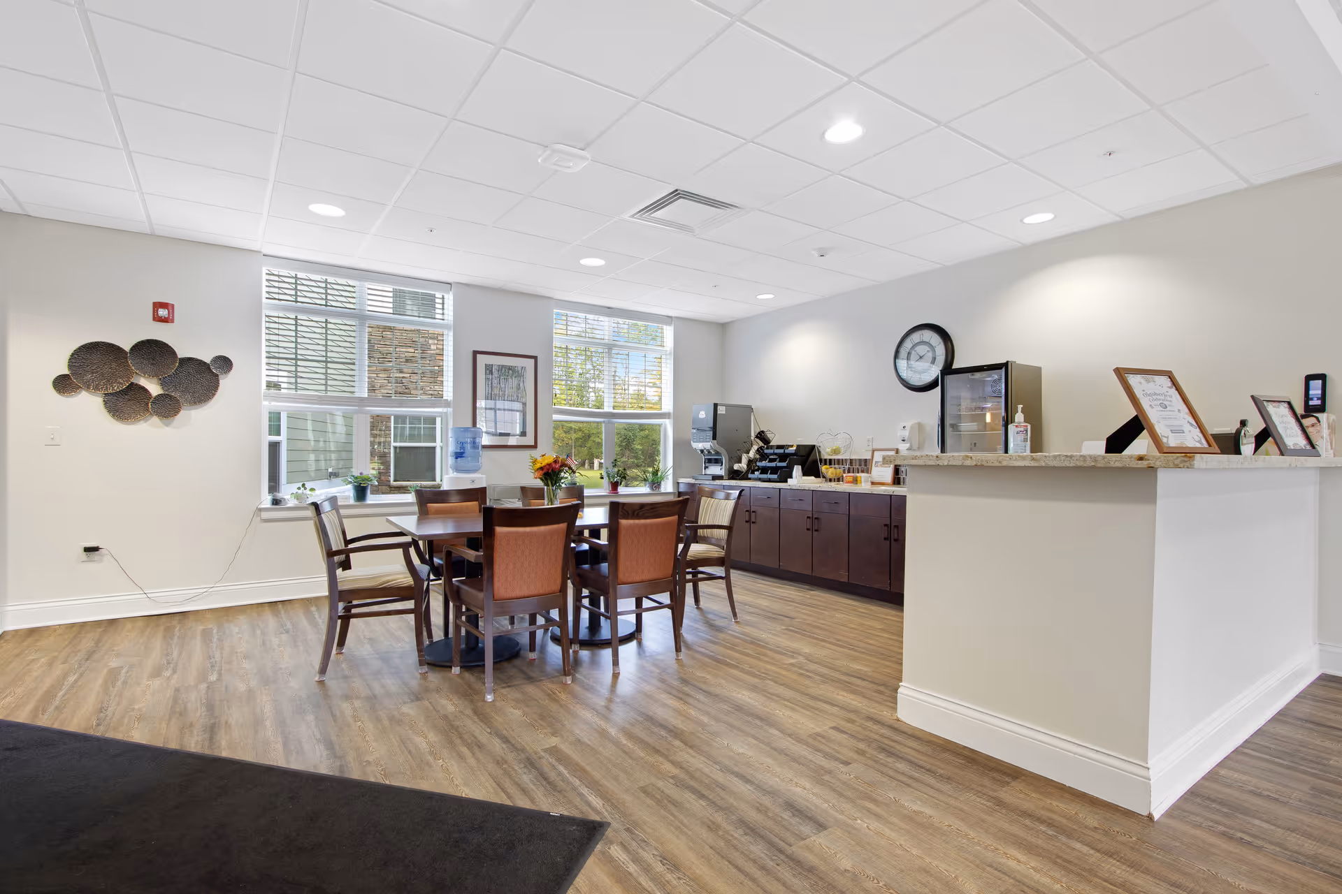 A bright and clean common area with a round table surrounded by six chairs in the center. The room has large windows letting in natural light, a water dispenser, and a counter with a coffee machine and other refreshments. The floor is wood, and the walls are decorated with a circular wall art and framed pictures. A clock is mounted on the wall above the counter.