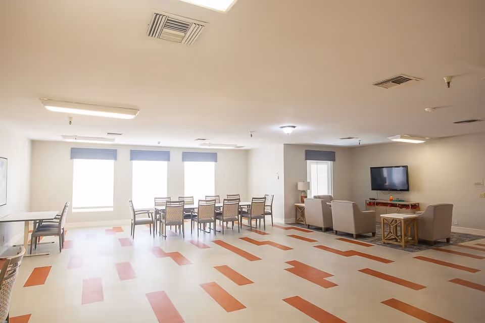 Bright communal room with dining tables and chairs on the left and a TV lounge area with armchairs on the right.