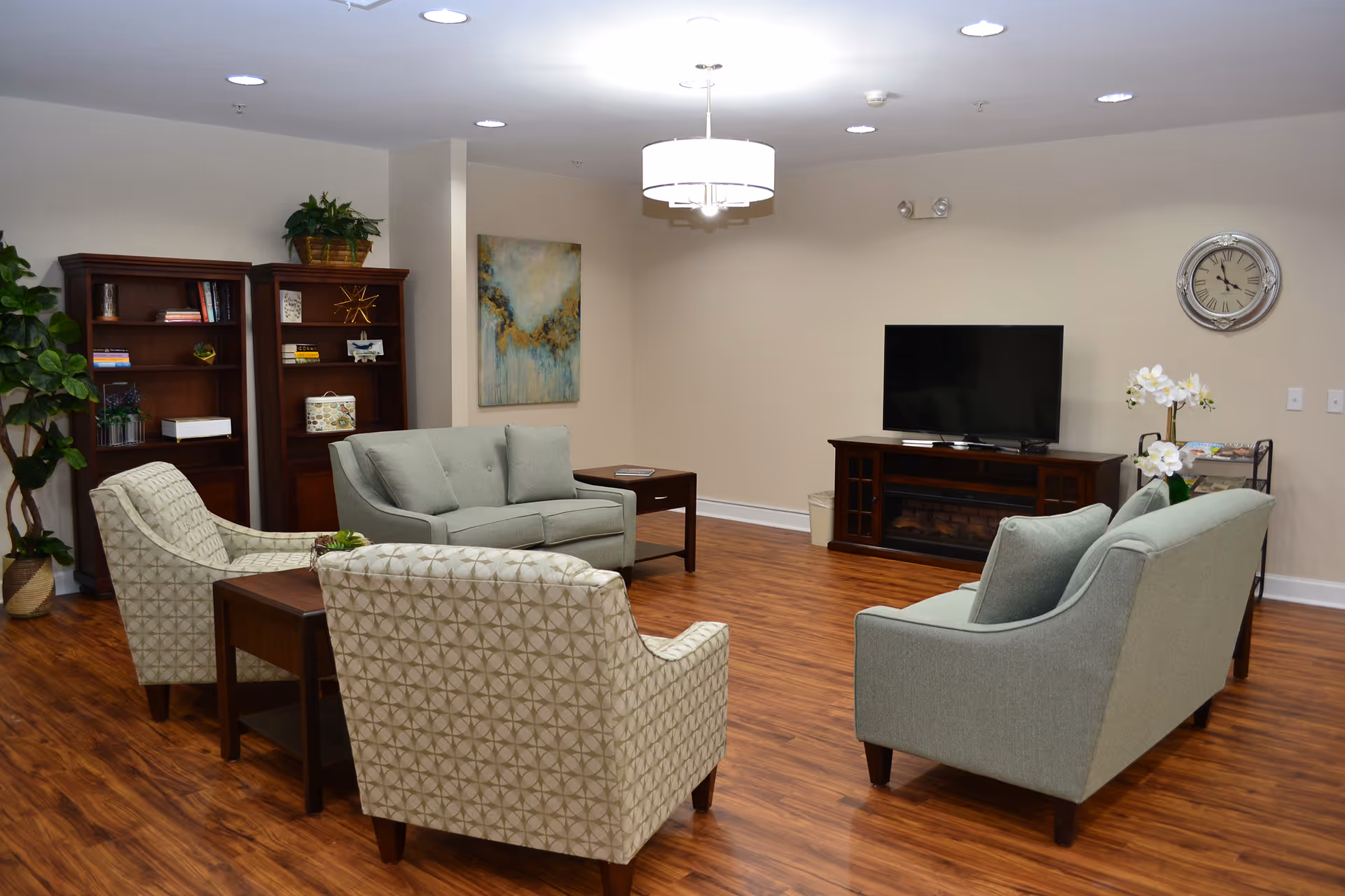 A cozy living room area with wooden flooring, two light green sofas, two patterned armchairs, a wooden coffee table, a TV on a wooden stand, a wall clock, bookshelves with books and decorative items, a potted plant, and a modern ceiling light fixture.